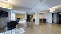 This contemporary community room is designed to accommodate a variety of social and recreational activities within a sleek, modern aesthetic. The space features an open floor plan finished with light hardwood flooring that enhances the bright, airy atmosphere created by generous natural light streaming through large windows.

The room is organized into several functional zones. A central gathering area offers round high-top tables paired with black bar-height chairs, providing flexible seating for casual mingling or group meetings. Recessed lighting throughout the ceiling ensures the space is well-lit at all hours.

A fully equipped kitchenette lines one wall, featuring white cabinetry, a mosaic tile backsplash, a beverage refrigerator, and small appliances — making it convenient for catering or self-service refreshments. Adjacent to this, a dedicated bar counter with additional bar seating offers a more lounge-like setting, also accented with a tile backsplash and pendant lighting.

An accent wall featuring a textured, patterned wallcovering in warm gold tones adds visual interest and warmth to the otherwise clean, neutral palette of white walls and structural columns. The overall design is polished and functional, making this community room a well-appointed space for events, meetings, and everyday social interaction.