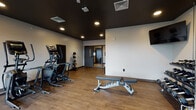 This fitness center is designed with a clean, modern aesthetic, combining crisp white walls with a bold dark ceiling that creates a focused and energizing workout environment. The space is finished with durable wood-look flooring throughout, providing both visual warmth and practical functionality underfoot.

The facility is comprehensively outfitted to support a wide range of training styles. Cardio equipment includes multiple elliptical trainers and at least one upright cardio machine, arranged to make efficient use of the floor space. For strength training, the room features a multi-functional cable machine, a full rack of dumbbells spanning a broad weight range, and adjustable and flat workout benches. A dedicated medicine ball rack holds a tiered selection of weighted balls for functional fitness exercises.

Natural light enters through a large window on one wall, offering views to the exterior and contributing to the room's openness. Recessed ceiling lights supplement the natural light, ensuring consistent illumination throughout the space. Multiple wall-mounted flat-screen televisions are positioned at viewing height throughout the room, allowing users to stay entertained during workouts.

Access to an adjacent room or corridor is provided through glass-paneled doors, adding transparency and connectivity within the facility. The overall layout is open and thoughtfully organized, giving users ample room to move freely between equipment.