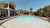 This outdoor pool area offers a refined, resort-like atmosphere highlighted by several distinctive architectural and decorative elements. The centerpiece is a generously sized swimming pool with clear turquoise water, framed by a brick-bordered deck lined with wicker and sling-style lounge chairs ideal for sunbathing. Towering columnar supports and lush landscaping, including a mature palm tree and manicured evergreens, enhance the upscale ambiance.

A standout feature is the ornate Victorian-style iron pavilion, painted in sage green, with intricate scrollwork panels, arched transom details, and a vaulted ceiling. Inside, wicker seating arrangements provide a shaded retreat with direct views of the pool. The open ironwork walls allow natural light and outdoor breezes to flow through freely.

Adjacent to the pool area, a covered outdoor kitchen is equipped with multiple full-sized gas grills beneath string lighting, offering a dedicated space for outdoor entertaining. A sculptural stone fountain labeled "Fontaine Bleu," adorned with detailed figural reliefs, serves as a striking decorative focal point within the landscaped grounds. The surrounding brick residential buildings complete the cohesive, elegantly designed outdoor environment.