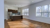This single-floor space showcases a thoughtfully designed open-concept layout that seamlessly connects the kitchen and living areas. The rich hardwood flooring runs throughout, catching natural light that pours in through multiple large windows fitted with horizontal blinds, creating a warm, sun-drenched atmosphere.

The kitchen is a standout feature, outfitted with deep espresso-toned cabinetry that provides ample storage and a striking visual contrast against the light-toned walls. A central island with a light stone countertop serves as both a prep space and a casual gathering point, anchored beneath a pair of modern pendant lights that add a contemporary touch. A stainless steel range with a sleek overhead hood and a stainless steel refrigerator complete the well-appointed culinary space. A subway tile backsplash in cool grey tones adds texture and refinement to the kitchen's backdrop.

The adjoining living area is open and expansive, with clean white walls and recessed lighting contributing to a bright, minimalist aesthetic. The abundance of natural light and the seamless flow between spaces create an inviting, airy ambiance well-suited for both everyday living and entertaining.