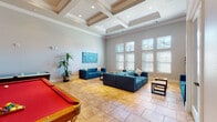 This single-floor recreational space is designed for comfort and entertainment, combining thoughtful architectural details with a welcoming, open atmosphere. The main lounge area is anchored by striking coffered ceilings with recessed lighting, adding depth and elegance to the room's generous vertical height. Warm-toned large-format tile flooring runs throughout, providing a cohesive and easy-to-maintain surface underfoot.

Ample natural light streams in through multiple tall, double-hung windows fitted with white plantation shutters, offering both brightness and privacy. The seating area features plush teal sectional and sofa sets arranged around low wooden coffee tables, creating comfortable conversation groupings. Vibrant wall art in blues and teals enhances the cohesive color palette throughout the space.

The game room section includes a classic pool table with a bold red felt surface and an air hockey table, making it an ideal gathering spot for recreation. A wall-mounted flat-screen TV adds to the entertainment options. Adjacent areas reveal a striking stone accent wall surrounding a fireplace, lending a rustic contrast to the otherwise modern interior. Lush potted tropical plants are placed throughout, bringing touches of greenery and life to the space. Overall, the layout flows smoothly between lounging and activity zones, making this a versatile and inviting communal area.