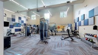 This expansive fitness center is thoughtfully designed to accommodate a wide range of workout styles and fitness goals. The main gym area impresses with soaring ceilings and clerestory windows that allow natural light to fill the space, complemented by sleek linear LED lighting overhead. The flooring throughout features a distinctive herringbone-patterned carpet tile, adding a modern aesthetic to the environment. Full-length mirrors line the walls, enhancing the sense of openness while providing functional visibility during workouts.

The main floor is extensively equipped with selectorized strength machines, cable systems, free weights including a full dumbbell rack and kettlebell selection, adjustable benches, and a multi-functional rig complete with a heavy bag, TRX suspension trainers, and battle accessories. Acoustic panels in blue and gray tones are mounted on the walls, managing sound effectively throughout the space.

A dedicated cardio area houses a row of treadmills positioned near windows, while a separate cycling studio offers a clean, light-filled environment with smooth wood-look flooring, spin bikes, wall-to-wall mirrors, dual wall-mounted televisions, and decorative mosaic tile art panels. A yoga mat storage nook and open floor space support stretching and group fitness activities, rounding out this comprehensive facility.
