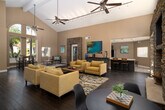 Located at 2500 Sea Cliff Way in Oceanside, California, this single-floor community space offers an open, welcoming atmosphere defined by impressive architectural details and thoughtful design. The centerpiece of the main lounge is a dramatic vaulted ceiling with exposed beams, ceiling fans, and track lighting that illuminates rich dark hardwood floors throughout. A floor-to-ceiling stacked-stone accent wall frames a built-in fireplace and mounted television, creating a striking focal point. Arched transom windows flood the space with abundant natural light while framing views of surrounding palm trees and outdoor amenities. French doors provide seamless access to the exterior. A dining area with a modern round table and a casual high-top dining set offer flexible seating options, while a stacked-stone archway leads to the office and additional rooms. The well-appointed kitchen features dark wood cabinetry with a built-in wine rack, light countertops, stainless steel appliances, and tile flooring, with a glass door opening directly to a pool area. The private office is finished with recessed lighting, hardwood floors, and ample natural light from multiple windows, making it a functional and professional workspace. The warm, neutral color palette and curated artwork contribute to a cohesive, inviting ambiance throughout.
