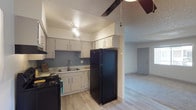 This single-floor space offers a functional and well-appointed layout designed for comfortable everyday living. The kitchen serves as a central feature, outfitted with white cabinetry that extends to the upper walls for generous storage capacity. A distinctive blue-gray subway tile backsplash adds a modern touch, complementing the clean lines of the white countertops and double basin sink. Black appliances, including a full-size refrigerator and a range, create a bold contrast against the lighter cabinetry and walls. Wood-look flooring runs throughout the kitchen, lending warmth and continuity to the space. A ceiling fan is positioned overhead, providing both air circulation and a subtle design element. The kitchen opens directly into a carpeted living area, creating a seamless flow between the two spaces and enhancing the overall sense of openness. A hallway visible from the kitchen leads further into the unit, connecting to additional rooms. The textured ceiling adds a classic touch throughout, while the overall layout maximizes the use of available space, making this a practical and inviting single-floor residence.