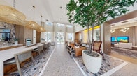 Located on East Sunrise Boulevard in Fort Lauderdale, this thoughtfully designed single-floor amenity space offers an impressive blend of coastal sophistication and modern functionality across a generously sized layout. Soaring double-height ceilings flood the interior with natural light through expansive windows dressed in sheer linen drapery, creating an airy and refined ambiance throughout.

The open-plan kitchen, dining, and living area serves as the heart of the space. The gourmet kitchen showcases warm wood cabinetry, a dramatic mosaic tile backsplash, stainless steel appliances, and a substantial marble-topped island with bar seating — all illuminated by sculptural woven rattan pendant lights. The adjacent living area features plush seating, statement indoor trees in oversized planters, and a slatted wood accent wall anchored by a large flat-screen display.

A secondary lounge area continues the coastal-modern aesthetic with layered textiles, botanical accents, and curated décor. Colorful geometric terrazzo flooring ties the communal areas together with a distinctive design flair.

The spacious primary bedroom provides a private retreat, while a dedicated indoor golf simulator room adds a unique recreational dimension to the space. Together, these elements deliver a well-rounded, amenity-rich environment that reflects the vibrant lifestyle of South Florida.