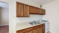Located at 1445 E Broad St in Columbus, Ohio, this single-floor unit offers a practical and comfortable layout across several well-defined living spaces. The kitchen is equipped with warm-toned wood cabinetry extending to the upper walls, offering generous storage space. Light-colored countertops provide ample workspace, complemented by a double stainless steel sink and a white gas range, while light tile flooring keeps the area clean and easy to maintain. An entry area features the same tile flooring and a window fitted with blinds, providing a welcoming transition into the home. The living spaces are finished with wall-to-wall carpeting in a neutral tone, creating a comfortable setting. One carpeted room benefits from dual windows that allow natural light to fill the space from multiple angles. Another carpeted room includes a closet and an adjoining passage, adding to the functional flow of the layout. The bathroom features classic white square tile surrounding the bathtub and shower area, with a window fitted with blinds that introduces natural light. A parquet-style floor and a baseboard heater complete the bathroom's traditional character. Throughout the unit, neutral wall tones and clean white trim contribute to a bright and cohesive aesthetic.