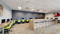 Located at 2508 SE Delaware St in Minneapolis, MN, this single-floor workspace is a well-appointed computer lab and collaborative hub designed for productivity and versatility. The open-plan layout accommodates a generous number of workstations arranged along perimeter desks lining a bold navy blue accent wall, each equipped with desktop computers and vibrant lime-green ergonomic chairs that add a lively pop of color to the environment.

At the center of the room stands a substantial wood-finished bar-height island with sleek black adjustable stools on both sides, serving as a flexible collaborative or casual work surface with built-in power access points. The flooring throughout consists of patterned commercial carpet tiles in neutral tones, contributing to a professional yet approachable atmosphere.

Natural light flows into the space through a row of large windows and glass-paneled double doors along one wall, offering views of a covered outdoor common area beyond. The drop ceiling features recessed and flush-mounted fluorescent panel lighting, ensuring consistent, even illumination throughout the room. Colorful abstract artwork adorns the walls, adding a creative touch to the functional environment. The space is efficiently organized to support both individual focused work and group collaboration.