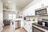 This single-floor unit offers a well-designed, open-concept layout that maximizes functionality within a compact footprint. Light wood-tone flooring runs consistently throughout, contributing to a bright, cohesive aesthetic. Neutral gray and white walls keep the space feeling airy and clean.

The kitchen is outfitted with white shaker-style cabinetry featuring matte black hardware, light marble-style countertops, and a full suite of stainless steel appliances including a range, over-the-range microwave, dishwasher, and refrigerator. Pendant lighting above the peninsula adds a contemporary touch, while the open sightlines connect the kitchen directly to the living area.

The living and sleeping area is combined within a single open space, with large windows dressed in horizontal blinds allowing substantial natural light. A ceiling fan overhead adds comfort and practicality.

The bathroom features a single-sink vanity with white cabinetry, a large mirror, white countertops, and a full tub-shower combination with a curtain rod. Light wood-tone flooring continues here as well.

The entryway is clean and functional, with a modern front door, an adjacent interior door, and neutral walls that set a welcoming tone upon entry.