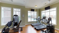 The fitness center is a bright, single-level space designed to support a well-rounded workout routine. The room is finished with warm hardwood-style flooring and sage-green walls accented by crisp white crown molding and baseboards, creating a clean and inviting atmosphere. Large multi-pane windows line the walls, flooding the space with abundant natural light and offering pleasant views of the outdoor pool area and surrounding grounds, making workouts more enjoyable. A wall-mounted flat-screen television is positioned near the windows, providing entertainment during exercise sessions. The cardio section is well-appointed with multiple treadmills, including models featuring integrated digital displays for tracking workout metrics. A cable resistance strength training machine is positioned along the side wall, offering users options for targeted muscle conditioning. The layout of the equipment allows for comfortable movement throughout the space without feeling cramped. Ventilation is supported by ceiling vents, ensuring air circulation during use. Overall, the fitness center offers a functional and pleasant environment suited for both cardio and strength-focused workouts.