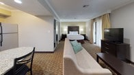 Located on Canada Ave in Orlando's Dr. Phillips neighborhood, this single-floor suite offers a well-rounded layout designed for comfort and extended-stay functionality. The open living and sleeping area is generously proportioned, featuring patterned carpeting, crown molding, and large sliding glass doors that invite natural light and provide views of the surrounding greenery. A queen bed with a dark wood headboard anchors the sleeping area, while a comfortable sofa and armchair with teal accent pillows define the separate sitting space. A standout feature of the living area is the white-surround fireplace, paired with framed artwork above, lending warmth and a residential feel. A flat-screen television on a dark wood media console completes the entertainment setup. The full kitchen is equipped with stainless steel appliances — including a full-size refrigerator, dishwasher, microwave, and range — set against dark cabinetry and granite countertops with hardwood-style flooring. A dining table provides space for meals. The bathroom includes a granite-topped vanity with ample counter space, a separate water closet with a combination tub and shower, and grab bars for accessibility. Wood-look flooring and clean, neutral finishes complete the space.