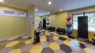This energetic yoga and fitness room is designed to inspire movement and wellness. The space is immediately defined by its striking hexagonal tile flooring, rendered in a bold palette of yellow, grey, black, and white — a playful geometric pattern that sets a lively, motivating tone throughout.

The walls are painted in a fresh, light green that enhances the room's bright and energizing atmosphere. Recessed lighting in the ceiling provides even, consistent illumination across the open floor plan, which offers ample room for stretching, yoga, and group exercise sessions.

One wall is adorned with a vibrant botanical-patterned wallcovering in coordinating greens and yellows, serving as a focal point backdrop for a wall-mounted flat-screen television — ideal for streaming guided workouts or fitness classes. A large mirror wall in the exercise area reflects the full space, enhancing the sense of openness and allowing users to monitor their form during workouts.

The room is well-equipped with a variety of fitness tools, including medicine balls on a rack, stability balls in multiple sizes, kettlebells, colorful dumbbells, and a BOSU balance trainer. Windows along one wall offer natural light and views of an outdoor pool area, connecting the interior to the surrounding environment. Multiple doorways provide convenient access to adjacent rooms and facilities.