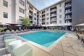 Located at 385 N Angier Ave NE in Atlanta's vibrant Old Fourth Ward neighborhood, this outdoor amenity space delivers a resort-style experience in the heart of the city. The centerpiece is a generously sized, crystal-clear swimming pool with lap lanes, framed by a wide, well-appointed sun deck finished in large-format concrete pavers. Rows of chaise lounges and lounge chairs line the pool's perimeter, providing ample space for relaxation and sunbathing. Shaded seating areas with market umbrellas and pergola structures offer comfortable retreats from the sun, while clusters of modern cube seating add flexible social gathering spots throughout the deck. The pool area is thoughtfully enclosed by a multi-story residential building featuring balconied units, warm wood accents, and a contemporary facade that creates a sense of intimate privacy. At the same time, open sightlines toward the Atlanta skyline — including views of notable high-rise towers — lend the space a distinctly urban energy. Mature trees and greenery soften the hardscape, enhancing the overall ambiance. Ground-floor amenity spaces visible through glass storefronts connect seamlessly to the pool deck, reinforcing the cohesive indoor-outdoor design. This space is ideal for leisure and community, combining modern design with a vibrant Atlanta backdrop.