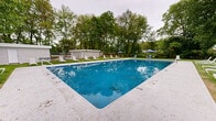 Located at 98 Robert Treat Dr in Milford, Connecticut, this outdoor recreational area offers a well-maintained community pool and grounds designed for relaxation and leisure. The centerpiece of the space is a generously sized rectangular swimming pool with a clean concrete surround, depth markers visible along the edges, and vivid blue water that reflects the surrounding landscape. The pool is equipped with entry steps and is bordered by a wide pool deck that provides ample room for poolside activities.

Surrounding the pool is a sprawling, lush green lawn that extends in multiple directions, framed by tall, mature trees that provide natural shade and a sense of seclusion. Scattered throughout the lawn and pool deck are white lounge chairs and chaise lounges, along with patio chairs and blue market umbrellas, creating comfortable spots for sunbathing and outdoor relaxation.

Two low-profile utility or support structures with light-colored siding are positioned at the rear of the property, likely serving as storage or equipment facilities. A chain-link fence borders portions of the perimeter, with views of a neighboring multi-story brick residential building visible beyond. The overall ambiance is that of a peaceful, tree-lined outdoor retreat, well-suited for warm-weather recreation within a residential community setting.