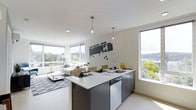 This single-floor space showcases a thoughtfully designed open-concept layout that seamlessly integrates the kitchen, dining, and living areas into one cohesive environment. The kitchen is anchored by a generous island with a crisp white countertop and dark charcoal cabinetry, providing both prep space and casual seating. Stainless steel appliances, including a dishwasher, and a modern basin sink with a contemporary arc faucet complete the functional kitchen setup. Two slender pendant lights hang above the island, adding a warm, stylish accent to the neutral palette.

The living area flows naturally from the kitchen, furnished with a light-toned sofa and a sleek glass coffee table atop a textured area rug. A decorative keyboard-inspired wall graphic serves as a creative focal point, lending the space a modern, artistic character. Large floor-to-ceiling windows flood the interior with abundant natural light and frame sweeping views of lush treetops and a distant waterway. Light hardwood flooring and white walls contribute to the bright, airy ambiance throughout, creating a clean and contemporary living environment.