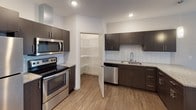Unit 310 is a well-appointed, single-floor residence designed with a contemporary aesthetic and functional layout. The kitchen serves as a standout feature, outfitted with dark espresso cabinetry, granite countertops, a white subway tile backsplash, and a full suite of stainless steel appliances including a refrigerator, electric range, microwave, and dishwasher. A dedicated walk-in pantry provides generous storage directly off the kitchen. Wood-look flooring runs throughout the kitchen and entry areas, transitioning to plush carpet in the living and bedroom spaces. The open living area is bathed in natural light from a sliding glass door that leads to a private balcony, offering outdoor views. Exposed ductwork adds an industrial design accent carried through the bedrooms as well. Each bedroom features a ceiling fan, carpeted floors, and a window with exterior views. The primary bathroom includes a dual-sink vanity with granite countertops, dark cabinetry, and direct access to a walk-in closet. A second full bathroom mirrors the same refined finishes with a double vanity. The dedicated laundry room houses a full-size front-loading washer and dryer alongside overhead wire shelving for added convenience.