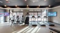This fitness center is designed to support a full range of workout routines within a thoughtfully organized, single-level space. The interior is finished with a dark ceiling accented by recessed lighting, which contrasts with light-toned walls and carpeted flooring to create a modern, energizing atmosphere.

The cardio zone is positioned along a wall of expansive floor-to-ceiling windows, allowing abundant natural light to fill the space while offering views of the surrounding exterior. This area houses multiple treadmills, elliptical machines, and a stair climber, all arranged to take advantage of the outdoor sightlines.

The strength training area features a multi-station cable machine in red, a dedicated free weight section with a full dumbbell rack, adjustable benches, and a heavy punching bag for boxing-style training. A large wall mirror enhances the sense of space and allows users to monitor their form throughout their workout.

An accent wall displaying motivational words and athletic imagery adds visual energy to the space, accompanied by multiple wall-mounted flat-screen televisions positioned throughout. The overall design balances functionality and aesthetic appeal, creating an inviting environment suited to a variety of fitness activities.