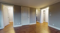 This single-floor space offers a well-appointed layout that balances classic architectural details with practical functionality. The main living area is generously proportioned, showcasing rich, dark-stained hardwood flooring that catches and reflects natural light from multiple double-pane windows. Crown molding runs along the ceiling perimeter, adding a refined touch to the neutral gray-toned walls. Baseboard heating units line the walls beneath the windows, ensuring consistent warmth throughout the space.

The layout includes multiple rooms connected by clean white-trimmed doorways. A secondary room features lighter hardwood flooring and its own window with views of the surrounding neighborhood. Both rooms benefit from an abundance of natural light, creating a bright and airy atmosphere.

A dedicated hallway leads to a closet and a full bathroom. The bathroom is notably well-finished, featuring floor-to-ceiling large-format stone-look tiles in warm gray tones lining the walk-in shower stall. Matching tile flooring ties the space together cohesively. Brushed nickel fixtures and a window add both style and ventilation to the room.

Overall, the space presents a clean, move-in-ready environment with consistent finishes, thoughtful storage solutions, and a comfortable flow between rooms.