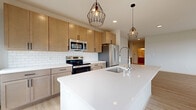 This contemporary single-floor residence showcases a cohesive modern design characterized by light wood-tone luxury vinyl plank flooring, crisp white walls, and clean architectural lines throughout. The open-concept kitchen serves as the unit's centerpiece, featuring a large white quartz waterfall island with an undermount sink and industrial-style faucet, light wood cabinetry, white subway tile backsplash, and stainless steel appliances including a range, microwave, and refrigerator. Two geometric cage-style pendant lights add a stylish contrast overhead. Large floor-to-ceiling windows flood the kitchen and living areas with natural light while offering outdoor views. The bedrooms are generously sized, with one room featuring a wall of black-framed windows and another with access to a dedicated walk-in closet and proximity to an in-unit stacked GE washer and dryer. Additional rooms include built-in cabinetry for storage utility. The bathroom is outfitted with a full soaking tub, shower combination, and wood-tone flooring. A hallway connects all areas efficiently, maintaining an open and airy ambiance throughout the unit.
