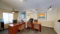 This single-floor professional office space is thoughtfully designed to balance functionality with a welcoming atmosphere. The entry area greets visitors with patterned carpeting, track lighting, and a pair of elegantly crafted wooden accent chairs with lattice detailing, set beside a small side table. Large glass double doors flood the entryway with natural light and offer views of a well-maintained outdoor courtyard. A pass-through window connects the reception zone to the adjacent interior, enhancing visibility and communication throughout the space. Crown molding and warm yellow-toned walls lend a polished, professional character throughout. The main work area features multiple rich wood-toned desks equipped with computer workstations, complemented by ergonomic seating and overhead ceiling fans for added comfort. Vibrant framed artwork depicting tropical and coastal scenes adds color and personality to the walls. Track lighting ensures consistent, adjustable illumination across all areas. A compact staff kitchen offers full functionality with white cabinetry, a gas range, microwave, refrigerator, dishwasher, and hardwood-style flooring, serving the practical needs of the team. Filing storage and a printer station round out the administrative amenities, making this a well-appointed and efficient professional environment.