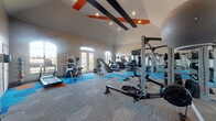 This fitness center is a thoughtfully designed, single-floor facility that offers a comprehensive workout environment. The space immediately stands out with its soaring vaulted ceilings, accented by bold diagonal beams painted in orange and black, lending the room a dynamic and energetic atmosphere. Large arched windows and French doors flood the interior with abundant natural light while offering views of the surrounding exterior.

The flooring features a striking pattern of hexagonal carpet tiles in shades of blue, orange, and gray, adding a modern visual identity to the space. The layout is generously proportioned, providing ample room to move freely between stations.

The cardio zone is well-stocked with treadmills, StairMaster steppers, elliptical trainers, and rowing machines, including an Endurance-brand rower. The strength training area includes an Octagon power rack, a cable machine, a dedicated dumbbell rack with a full range of weights, medicine balls, stability balls, and battle ropes. A heavy Everlast punching bag adds a functional fitness element. Floor-to-ceiling mirrors along one wall enhance the sense of space and allow for form monitoring during workouts.

A wall-mounted flat-screen television provides entertainment, while the overall design balances functionality with a vibrant, motivating aesthetic suitable for a wide range of fitness routines.