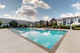 Located at 1100 Enterprise Way NW in Huntsville, Alabama, this outdoor amenity space centers around a generously sized, crystal-clear swimming pool designed for both relaxation and recreation. The pool features built-in in-water lounge chairs along its edges, inviting residents to unwind directly in the water, while a row of sleek slatted sun loungers lines the broad concrete pool deck on multiple sides, offering ample space for sunbathing. A white fabric gazebo provides a shaded retreat at one end of the deck, adding an elegant touch to the overall atmosphere. The pool area is fully enclosed by a black wrought-iron fence, ensuring both safety and a clean aesthetic boundary. Mature trees and manicured landscaping beds with fresh mulch are thoughtfully integrated around the perimeter, softening the hardscape and adding greenery. The surrounding multi-story residential buildings feature a modern farmhouse-inspired architectural style, with dark gray, navy, and white board-and-batten and siding exteriors, pitched rooflines, large windows, and private balconies that overlook the pool courtyard. The open-sky setting allows for abundant natural light throughout the day, creating a bright, resort-like ambiance that serves as a standout communal feature of this Huntsville property.