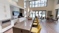 This contemporary amenity clubhouse is designed to impress, blending architectural drama with refined interior detailing across a single, well-appointed floor. The space is anchored by a striking lounge area featuring grand arched openings that frame sweeping views of the outdoor pool and courtyard beyond, flooding the interior with abundant natural light. Decorative lantern-style pendant lights hang above comfortable seating arrangements that include plush sofas and accent chairs, accented with sage green walls, textured wall art, and patterned rugs that add warmth and personality.

A spacious social hall showcases soaring double-height ceilings with expansive floor-to-ceiling windows and glass paneling, further enhancing the sense of openness. Wide-plank wood-look flooring runs throughout, complementing the clean, neutral palette. A fully equipped bar and prep area features sleek white cabinetry, a metallic tile backsplash, a wall-mounted flat-screen television, and a long polished counter lined with vibrant yellow upholstered bar chairs — an ideal setting for social gatherings.

An adjacent seating lounge offers a more intimate atmosphere, with carefully curated furnishings, accent lighting, and decorative wall installations that create a sophisticated yet inviting retreat. Throughout, the design seamlessly connects indoor and outdoor living, making this a standout communal amenity space.