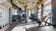 Located at 318 I St NE in Washington, DC's Capitol Hill neighborhood, this fitness center offers a thoughtfully designed, open workout space across a single floor. The layout is efficiently organized to accommodate a full range of exercise equipment without feeling cramped, making excellent use of its footprint.

Along the perimeter, floor-to-ceiling windows invite abundant natural light and provide views of the surrounding streetscape, creating an energizing atmosphere for workouts. The cardio zone features a row of modern treadmills with integrated screens, ellipticals, and stair climbers positioned directly in front of these expansive windows. A dedicated free weights area includes a full rack of dumbbells and adjustable workout benches, supporting a comprehensive strength training routine.

The design aesthetic is sleek and contemporary, with an exposed industrial-style ceiling fitted with track and pendant lighting that adds warmth to the dark, sophisticated color palette. A standout visual element is a striking installation of colorful iridescent panels near the entrance, adding an artistic flair to the space. Large mirrors along the walls enhance the sense of openness. Durable rubber flooring throughout ensures safety and comfort during exercise. The overall ambiance is polished and motivating, blending functional design with distinctive creative touches.