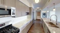 This contemporary single-floor residence showcases a thoughtfully designed open layout with high-end finishes throughout. The space features two distinct kitchen configurations: one with a generous island topped with granite countertops, pendant lighting, sleek taupe cabinetry, and stainless steel appliances including a gas range and refrigerator; and a second galley-style kitchen equally well-appointed with matching stone counters, tile backsplash, and an in-unit stacked washer and dryer nearby for added convenience.

The living area is bright and welcoming, finished with dark hardwood flooring, a ceiling fan, and dual sliding glass doors that open onto a private wraparound deck. This outdoor space features warm wood decking and elevated urban views of the surrounding cityscape. Multiple bedrooms are carpeted and filled with natural light through large windows overlooking the city. One bedroom connects directly to a primary bathroom featuring a wide vanity, tiled soaking tub, and an adjacent walk-in closet. A second full bathroom offers a tiled tub-shower combination with built-in shelving. The overall aesthetic is clean, modern, and move-in ready.