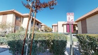 This outdoor community space is designed with pet owners in mind, offering a dedicated and well-maintained dog park area nestled within a residential complex of Southwestern-style buildings. The architecture throughout the surrounding complex is characterized by warm stucco exteriors, clay tile roofing, and earth-toned facades that reflect a classic desert regional aesthetic.

The dog park itself is enclosed by chain-link fencing, providing a secure environment for pets to roam freely. The ground surface features a combination of artificial turf and decomposed granite, offering both a clean play surface and low-maintenance landscaping suited to an arid climate. Pet agility equipment is thoughtfully placed within the space, encouraging active play and exercise for dogs of all sizes.

A clearly posted pet waste management station is situated at the entrance, emphasizing the community's commitment to cleanliness and responsible pet ownership. Mature desert-adapted trees and shrubs line the perimeter, providing light shade and a natural buffer. Under wide-open blue skies and ample sunlight, the space enjoys an inviting, open-air atmosphere. The overall setting reflects a community that prioritizes outdoor amenities and the well-being of its residents and their animal companions.