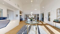 This contemporary single-floor community space makes an immediate impression with its dramatic double-height entry foyer, where a cascading gold pendant chandelier hangs above a herringbone-patterned hardwood floor and a glass-topped accent table. Floor-to-ceiling glass doors and transom windows flood the space with natural light, creating a bright and welcoming atmosphere throughout.

The open layout flows seamlessly from the grand entry into a spacious lounge area anchored by a linear gas fireplace framed by built-in white shelving units displaying curated décor. Comfortable seating — including plush sofas and vibrant orange accent chairs — surrounds the fireplace, while a separate social lounge features soaring clerestory windows, bold red seating, and direct access to the outdoors.

A well-appointed full kitchen serves the space with light wood cabinetry, dark countertops, stainless steel appliances, a glass-top island with bar seating, and a gold ring chandelier. A secondary beverage station offers additional prep space with a tile backsplash and open floating shelves.

The reception area features a striking angular desk with a brushed metal front panel, set against a backdrop of artwork and interior windows. A quiet corridor with a floor-to-ceiling built-in bookcase and pendant lighting provides a transitional gallery moment throughout this thoughtfully designed space.