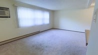 This single-floor space presents a straightforward and functional layout. The main room is finished with wall-to-wall carpet in a neutral beige tone, providing a soft underfoot feel that adds a sense of warmth to the interior. The room is well-lit, benefiting from a large picture window dressed with sheer vertical blinds that diffuse natural light evenly throughout the space, creating a bright and open atmosphere. The walls are finished in a clean, off-white tone that complements the light flooring and contributes to an airy feel. A baseboard heating unit runs along the exterior wall beneath the window, offering efficient and consistent climate control. The ceiling is smooth and neutral, maintaining the room's clean aesthetic. Dark baseboards provide a subtle contrast against the light walls, adding a modest defining detail to the overall design. The space is unfurnished, allowing for flexible arrangement and personalization. Overall, the room presents a calm, neutral environment well-suited for a variety of uses.