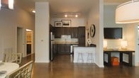 This single-floor space showcases a thoughtfully designed open-concept layout that seamlessly integrates the kitchen and living areas. The kitchen is a standout feature, outfitted with rich, dark wood cabinetry that extends to the upper walls, providing generous storage throughout. A classic white subway tile backsplash adds a clean, timeless contrast against the darker tones, while stainless steel appliances — including a refrigerator and dishwasher — lend a sleek, contemporary feel. The kitchen peninsula features a dark countertop and extends into a breakfast bar with white bar stools, offering a casual dining option that also enhances the open flow of the space. Track lighting overhead illuminates the kitchen efficiently, highlighting the functional design. The polished concrete floors run throughout, adding an industrial yet sophisticated character to the interior. Structural columns punctuate the open floor plan, subtly defining zones while maintaining the airy, connected feel of the space. The high ceilings amplify the sense of volume, contributing to the overall open and welcoming ambiance. Neutral white walls provide a versatile backdrop, allowing the dark cabinetry and concrete floors to serve as the defining design elements.