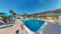 Located in Boca Raton, Florida, this outdoor amenity space centers around a generously sized swimming pool that serves as the focal point of the property. The pool area is enclosed by a white safety fence and finished with a clean concrete surround, offering ample space for sunbathing and relaxation. Multiple lounge chairs and vibrant teal umbrellas are arranged poolside, providing shade and comfort for guests enjoying the warm South Florida climate. Tall palm trees frame the background, adding a tropical ambiance characteristic of the region. The surrounding two-story building, finished in a warm yellow exterior with a dark red roof, wraps around the pool area in a U-shaped configuration, with balconies and large windows visible on the upper level. The architecture reflects a classic Florida mid-century resort style, with exterior corridors and railings on the upper floor. The open-air setting benefits from abundant natural sunlight and clear blue skies, creating a bright and inviting outdoor environment. This pool area functions as a shared outdoor amenity, offering residents or guests a comfortable retreat for leisure and recreation in a well-maintained, landscaped setting.