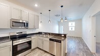 This efficiently designed single-floor unit offers a thoughtful layout across its four distinct spaces: a kitchen, bedroom, bathroom, and closet. The open-concept kitchen and living area serves as the heart of the unit, showcasing grey cabinetry with sleek hardware, dark granite countertops with a subtle tile backsplash, and a full suite of stainless steel appliances including a range, microwave, and dishwasher. A peninsula with a double sink anchors the kitchen, while stylish pendant lighting adds a modern touch. Wood-look flooring extends throughout the main living area, and a ceiling fan overhead adds both comfort and visual interest. Natural light filters in through a window near the front entry door, enhancing the open, airy feel of the shared living space. The bedroom offers a comfortable, carpeted retreat with a vaulted ceiling detail, a ceiling fan, and a window that brings in natural light and neighborhood views. The bathroom is clean and functional, featuring a full soaking tub with a shower and brushed nickel fixtures set against bright white walls. A dedicated closet area with laundry hookups and additional storage adds everyday practicality to this compact yet complete living space.