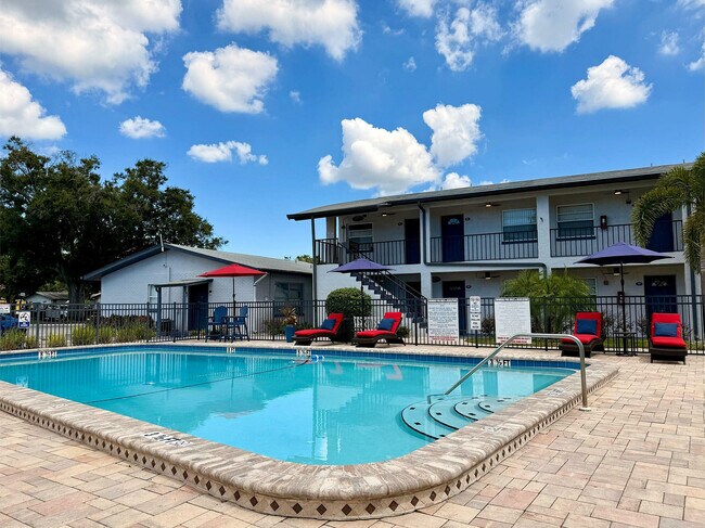 Encantador complejo de apartamentos con una piscina reluciente rodeada de tumbonas, sombrillas de colores vivos y exuberante vegetación bajo un cielo azul brillante. - Waterside Village