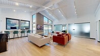 This thoughtfully designed single-floor space combines open-concept communal living with functional workspaces, making it an ideal gathering and productivity hub. The centerpiece of the space is a dramatic vaulted ceiling with exposed white-painted beams and tongue-and-groove paneling, soaring overhead to create a striking sense of volume and airiness. Track lighting runs along the ceiling lines, complementing the abundant natural light that pours in through large black-framed windows and a peaked clerestory window arrangement flanking a slatted wood accent wall with a wall-mounted flat-screen TV.

Light hardwood flooring flows seamlessly throughout, anchoring the open layout. The main lounge area features a generous seating arrangement with neutral sofas and rich terracotta-hued velvet armchairs, centered on a patterned area rug. A large ceiling fan enhances comfort in this airy zone.

Along one wall, a built-in counter with teal bar-height chairs provides a dedicated workspace with views to the outdoors. Additional work and utility amenities, including a printer station and water dispenser, are neatly tucked nearby beneath beamed ceilings. Frosted sliding glass doors reveal semi-private office or meeting rooms, offering flexibility while maintaining an open, light-filled aesthetic. A display shelving unit and tasteful artwork add polished finishing touches throughout.
