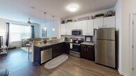 Located at 9200 University City Blvd in Charlotte, NC, this single-floor residence offers a thoughtfully designed layout suited for comfortable, functional living. The open-concept kitchen is a standout feature, outfitted with stainless steel appliances including a refrigerator, electric range, built-in microwave, and dishwasher. Two-tone cabinetry — white upper cabinets paired with dark lower cabinets — frames granite countertops, while warm wood-style flooring and pendant lighting add a modern, inviting touch. The kitchen flows seamlessly into the living area, which benefits from natural light through large windows fitted with blinds and drape panels. A warm tan leather sectional sofa and vibrant abstract wall art contribute to the welcoming ambiance of the living space. Each bedroom is designed with functionality in mind, featuring built-in headboard shelving units that incorporate display shelves and an integrated desk — ideal for studying or working from home. Ceiling fans and large windows with views of the surrounding area enhance comfort and brightness. The bathrooms are finished with wood-style flooring, tub/shower combinations, and clean, neutral finishes. Overall, this residence combines practical design elements with a warm, livable aesthetic throughout every room.