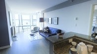 This single-floor space showcases a contemporary open-concept design that effortlessly connects the kitchen and living areas. The kitchen is anchored by a substantial granite-topped island or bar counter with rich, dark veining, providing both a prep surface and casual seating space. The living area is furnished with a tufted leather sofa, a sculptural accent chair, and a glass coffee table, all arranged on a light area rug atop smooth hardwood-style flooring. Two framed artworks adorn the clean white walls, adding a polished, curated touch. A sleek floor lamp provides ambient lighting to complement the abundant natural light streaming in through expansive floor-to-ceiling windows. These large windows offer open views of the surrounding cityscape and provide access to what appears to be an outdoor balcony, enhancing the sense of space and airiness throughout. The overall ambiance is modern and refined, with a neutral color palette of whites, greys, and dark accents creating a cohesive, sophisticated aesthetic throughout the unit.