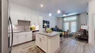 This single-floor residence offers a thoughtfully designed open-concept layout that seamlessly connects the kitchen, dining, and living areas. The kitchen is a standout feature, showcasing crisp white shaker cabinetry, granite countertops, a grey subway tile backsplash, and a full suite of stainless steel appliances. A central island with pendant globe lighting above serves as both a functional workspace and an informal gathering spot. Light wood-style flooring runs throughout the main living areas, enhancing the bright, airy feel of the space. The living room is generously proportioned, bathed in natural light from large windows dressed with blinds and sheer curtains, and is anchored by a ceiling fan overhead. The bedroom continues the warm, welcoming tone with plush carpeting, a large window that invites abundant natural light, and ample room for comfortable furnishings. The bathroom is well-appointed with white cabinetry, a granite vanity countertop, a large mirror, and stylish lighting fixtures, maintaining the cohesive design language found throughout the residence. Overall, the space presents a clean, contemporary aesthetic with high-quality finishes and a functional flow ideal for modern living.