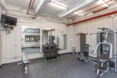 This fitness center on Walton Street NW in downtown Atlanta offers a well-equipped workout facility within a distinctive industrial-style space. The room features exposed painted brick walls, visible overhead ductwork and piping, and durable rubber-coated flooring designed to withstand heavy use. Large wall-mounted mirrors along one side visually expand the space and allow users to monitor their form during exercise. The facility is outfitted with a range of equipment to support varied workout routines, including a multi-station cable and strength machine, a treadmill, an elliptical trainer, and a full rack of dumbbells paired with a flat weight bench. Overhead industrial lighting ensures the space is well-lit throughout. Entry is provided through glass-paneled double doors, and posted fitness center rules indicate a managed, community-oriented environment. Windows along the exterior walls allow for natural light to supplement the overhead fixtures. The overall ambiance is functional and utilitarian, reflecting the building's urban character while providing residents or occupants with convenient access to essential fitness amenities in the heart of Atlanta.