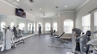 This fitness center is thoughtfully designed to support a well-rounded workout experience. The room features a distinctive curved wall lined with elegant arched windows that allow abundant natural light to fill the space, creating a bright and energizing atmosphere. Crown molding along the upper walls adds a refined architectural touch, complementing the clean, neutral color palette throughout.

The flooring consists of durable rubber gym matting, providing both safety and functionality for a range of exercises. A large wall-mounted mirror spans one side of the room, enhancing the sense of openness while allowing users to monitor their form during workouts. Recessed lighting and a ceiling fan ensure the space remains well-lit and comfortable during use.

The facility is outfitted with a selection of strength training equipment, including cable machines, adjustable weight benches, and free weights, catering to users with varying fitness goals. A wall-mounted flat-screen television adds an entertainment element to the workout environment. The overall ambiance is clean, spacious, and well-organized, reflecting a commitment to providing a functional and welcoming fitness experience.