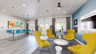 Located at 10455 E Via Linda in Scottsdale, Arizona, this single-story community clubhouse is designed to serve as a welcoming social and amenity hub. The entrance greets visitors with a dramatic vaulted ceiling, a statement chandelier, light wood-look flooring, and large glass doors that flood the space with natural light.

The main lounge area is a standout feature, showcasing bold teal accent walls, bold yellow high-back accent chairs arranged around a modern white coffee table, and a wall-mounted flat-screen TV — creating a comfortable yet visually striking gathering space. Patterned drape panels and recessed lighting add warmth and texture throughout.

Adjacent to the lounge is a coffee bar station, outfitted with a Keurig coffee machine, pod organizer, stainless steel mini-fridge, and white cabinetry — a convenient amenity for daily use. A high-top bar seating area with teal chairs offers an additional casual lounge option.

The fitness center is well-equipped with a cable weight machine, elliptical, weight bench, and other exercise equipment set on durable carpet tile flooring, with French doors opening directly to the outdoor pool area.

Outside, a covered patio provides shaded seating with wicker furniture and blue cushions, a ceiling fan, and a vending machine — ideal for relaxing in the Arizona sunshine. The overall design balances contemporary style with functional community amenities.