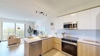Located at 1740 S Young Circle in Hollywood, Florida, this thoughtfully designed studio offers a bright and contemporary living experience on a single floor. The open-plan kitchen and living area serves as the heart of the space, featuring light wood-look flooring that runs seamlessly throughout. The kitchen is outfitted with stainless steel appliances, including a full-size refrigerator, electric range, and over-the-range microwave. Two-tone cabinetry — combining white upper cabinets with warm wood-grain lower units — is complemented by a herringbone tile backsplash and a peninsula with bar seating for three, making it ideal for both cooking and casual dining. The living area is bathed in natural light from a large sliding glass door that opens to a private balcony, offering sweeping views of the surrounding palm-lined landscape and the Hollywood skyline beyond. The bathroom continues the warm, modern aesthetic with a wood-grain vanity, white countertop, and an illuminated LED mirror. A dedicated walk-in closet provides generous storage with built-in wire shelving. The overall ambiance is clean, airy, and contemporary, making this an efficient yet stylish space in the heart of South Florida.