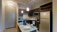 Located at 2000 Front Street in Summerville, South Carolina, this single-floor residence offers a thoughtfully designed living space with modern finishes throughout. The kitchen serves as a central highlight, showcasing rich dark cabinetry that provides ample storage, including an integrated wine rack for added functionality. Granite countertops extend across the workspace and onto a kitchen island, which doubles as a casual dining and entertaining surface. Stainless steel appliances — including a refrigerator, microwave, and range — lend a sleek, contemporary feel to the space. Overhead, a combination of pendant lighting and a track lighting system ensures the kitchen is well-lit and visually inviting. Light hardwood-style flooring flows through the area, complementing the warm tones of the cabinetry and stone surfaces. The layout is designed for both efficiency and comfort, with a practical workflow between the sink, cooking range, and refrigerator. The overall ambiance of the space is polished and modern, with carefully chosen fixtures and finishes that create a cohesive, upscale aesthetic suited to everyday living and entertaining alike.