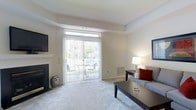 This single-floor space offers a welcoming and well-lit environment designed for comfortable everyday living. The living area serves as the centerpiece of the home, featuring soft carpeted flooring and a neutral color palette that creates a calm, airy atmosphere. A classic fireplace anchors one wall, adding a touch of warmth and character to the room. Opposite the fireplace, a large sliding glass door floods the space with natural light and provides direct access to an outdoor patio area, where greenery and open surroundings are visible. Vertical blinds offer flexible light control while maintaining the clean lines of the room. A transom window above the sliding door further enhances the brightness of the interior. The living space is thoughtfully laid out, accommodating seating, a coffee table, and additional furniture with ease, reflecting a practical yet comfortable floor plan. A wall-mounted television and built-in shelving add functional touches, while recessed ceiling details give the room subtle architectural interest. Overall, the space presents a tidy, move-in-ready environment with a light, neutral ambiance suitable for a variety of lifestyles.