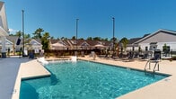 Located at 31 Quaker Ridge Drive in Carolina Shores, North Carolina, this two-floor community amenity center offers a well-rounded array of recreational facilities designed for active outdoor living. The centerpiece of the property is a sparkling resort-style swimming pool surrounded by a generous sun deck lined with lounge chairs, providing ample space for relaxation under the Carolina blue sky. Tall light poles illuminate the pool area for extended evening use, while the wrought iron perimeter fencing adds both security and visual appeal.

Adjacent to the pool, a dedicated pickleball and tennis court complex features a vibrant blue-and-green playing surface enclosed by chain-link fencing. The courts are equipped with regulation nets and overhead lighting, making them suitable for daytime and evening play. Lush landscaping with ornamental grasses, mulched beds, and young palm trees borders the courts, enhancing the coastal aesthetic of the community.

A bocce ball court runs alongside the main building, set within a well-maintained lawn area framed by neatly placed young trees. The clubhouse structure itself features classic craftsman-style architecture with board-and-batten siding, stone accents, gabled rooflines, and welcoming covered entry points. Together, these amenities create a vibrant, resort-inspired outdoor environment reflective of the Carolina Shores lifestyle.