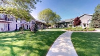 This residential community presents a thoughtfully designed exterior environment that immediately conveys a sense of care and quality. The grounds are defined by expansive, well-kept lawns that stretch across a central courtyard shared by multiple building wings. Curved concrete walkways guide residents through the property, flanked by meticulously maintained garden beds featuring ornamental grasses, flowering shrubs, and decorative mulched borders that add visual interest throughout the seasons.

Mature deciduous trees, including a striking red-leafed variety, provide natural shade and a sense of established character to the landscape. The buildings themselves feature a neutral brick and siding exterior with dark accent shutters, private balconies, and large windows that invite natural light into each unit. The multi-wing layout of the buildings creates an enclosed, community-oriented courtyard feel that enhances both privacy and a sense of neighborhood.

Under clear blue skies, the combination of vibrant greenery, thoughtful plantings, and clean architectural lines gives the property a bright and inviting ambiance. The overall setting reflects a commitment to exterior upkeep, offering residents an attractive and serene outdoor environment to enjoy year-round.