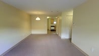 This two-floor space presents a clean, understated design characterized by light yellow-toned walls, smooth white ceilings, and wall-to-wall carpeting throughout the main living areas. The layout offers a generous open feel, with multiple rooms providing ample space for a variety of uses.

The living area is notably spacious, featuring neutral carpet flooring and large windows that allow natural light to filter in, brightening the interior. Baseboard heating runs along the perimeter of the rooms, ensuring efficient climate control, and a wall-mounted air conditioning unit is also present for year-round comfort.

A pendant light fixture marks the transition between the main living space and the kitchen area beyond, which is visible in the background and features wood-toned cabinetry, a window, and what appears to be a dishwasher. The kitchen connects smoothly to the living area, supporting an open and accessible flow between spaces.

An entry door with paneled detailing is visible near the kitchen, accompanied by an intercom panel, indicating a secured building entry system. Throughout, the space maintains a straightforward, functional aesthetic with neutral finishes that provide a versatile foundation for personalization.