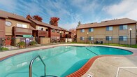Located at 7190 Calvine Rd in Sacramento, California, this outdoor amenity area serves as a central recreational hub within the residential community. The centerpiece of the space is a generously sized, freeform swimming pool with clear turquoise water, framed by a wide concrete pool deck and accented with a distinctive red brick coping border. Adjacent to the pool sits a circular spa, also finished with matching red and blue tile trim, providing a dedicated relaxation area.

A single-story clubhouse building anchors one side of the pool area, constructed with wood panel siding in warm tan and brown tones and topped with a sloped, shingled roof. The building houses utility and restroom facilities, with red-accented doors adding visual contrast. A covered overhang along the building's facade provides shaded space along the deck.

The entire pool area is enclosed by a black metal fence, ensuring security and privacy. Lush green lawn areas are visible beyond the fencing, and mature trees with autumn foliage provide a natural backdrop. The surrounding multi-story residential buildings, clad in wood siding with balconies and pitched rooflines, frame the space and contribute to the community's cohesive architectural character. The open layout and abundance of natural light make this an inviting outdoor gathering and recreation destination.