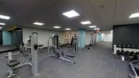 This fitness center is designed to provide a comprehensive workout environment within a single-floor layout. The space is outfitted with a range of professional-grade strength training machines, including cable and weight stack systems, strategically positioned to allow for efficient use of the floor space. The room features a dark, patterned carpet with subtle accent striping that defines the workout zones, while the neutral-toned walls are accented by a standout teal structural column that adds a pop of color to the interior. The ceiling is finished with a dark drop-tile grid and fitted with bright, recessed square panel lighting that evenly illuminates the entire area. An open corridor extends toward the back of the space, hinting at additional functional areas or utility rooms beyond. Free weights and supplementary equipment are also visible, rounding out the strength training offerings. The overall ambiance is clean, functional, and professional, reflecting a thoughtfully designed facility intended to support a variety of fitness routines in a spacious, well-lit environment.